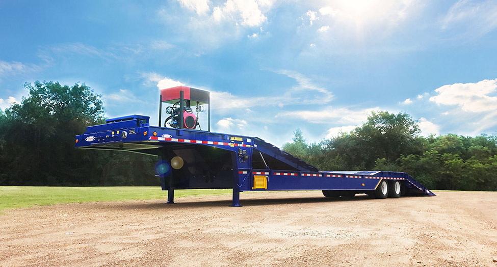 Blue Ledwell Hydratail Trailer with tail in down position. Red and black Self Contained Unit sitting on front deck of trailer. Sunny blue sky with green trees in the background and light brown dirt on ground.
